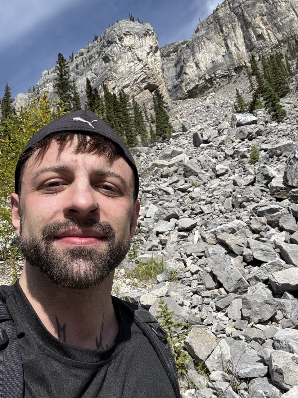 Demetri smiling on a mountain hike, with limestone cliffs and evergreens rising behind him